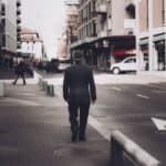back view of a man wearing a black blazer and a cap walking on sidewalk