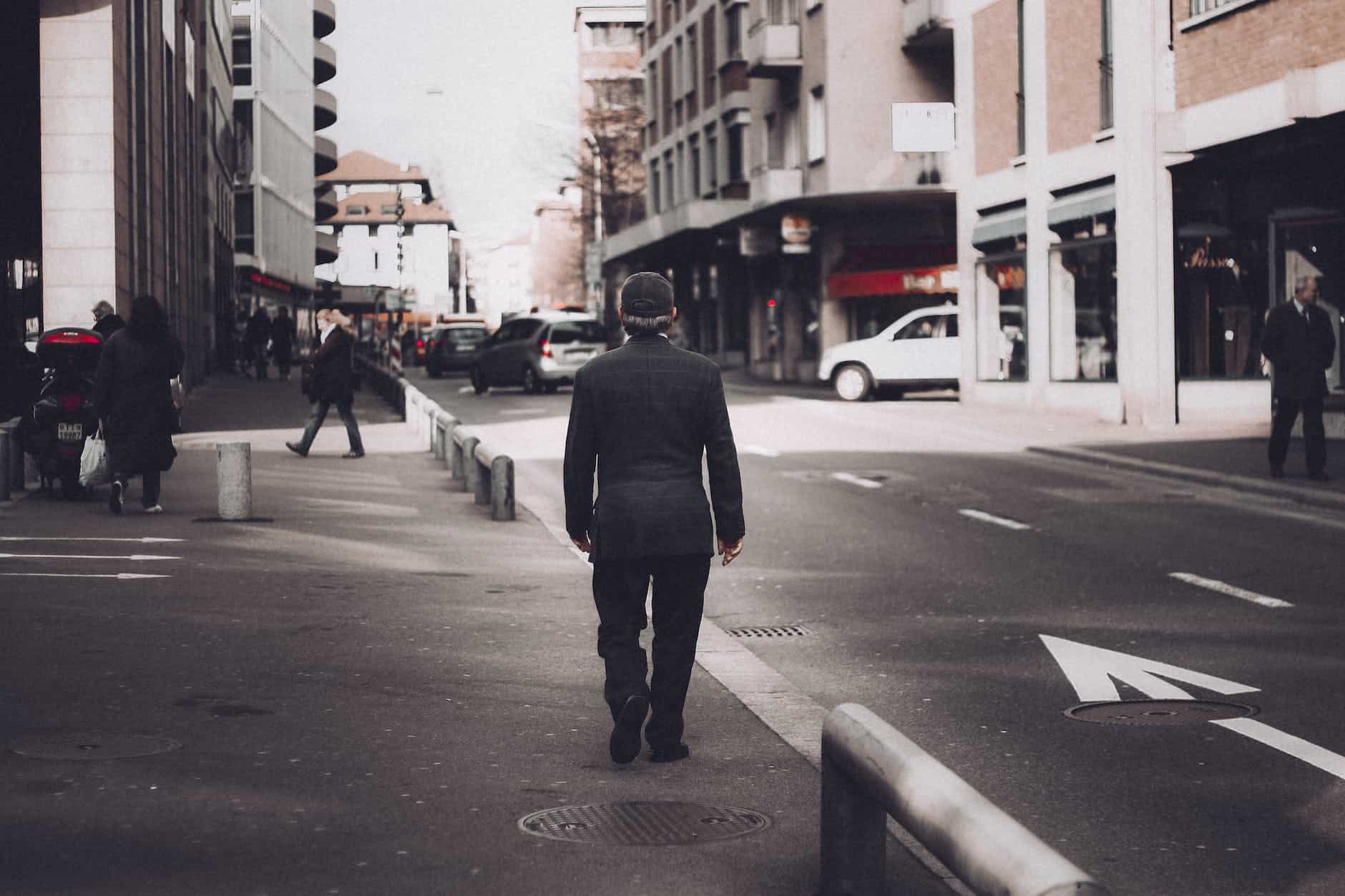 back view of a man wearing a black blazer and a cap walking on sidewalk